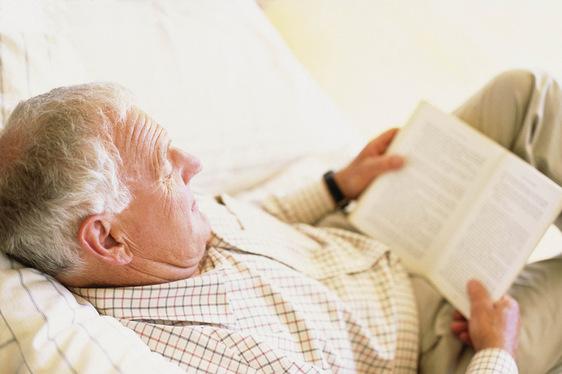 Älterer Mann liest entspannt ein Buch, während er in einem Bett mit weißen Laken liegt.